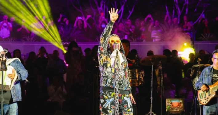 Ángel Pedraza of Grupo Kual with his arms up performing in downtown Los Angeles with lights in background.